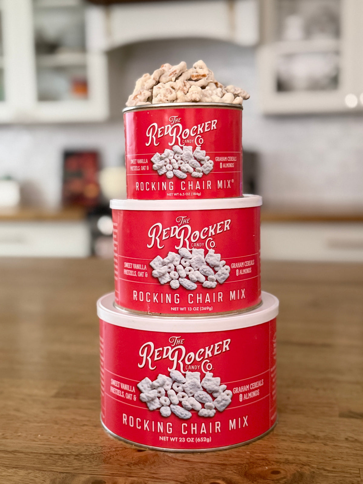 Three stacked cans of Red Rocker 'Rocking Chair Mix' snack mix on a wooden countertop in a blurred kitchen setting.